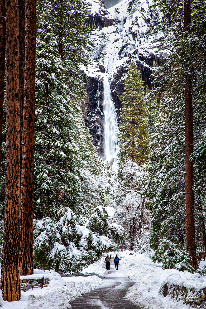 Two hikers are looking up from the path at the majestic winter beauty of Lower and Upper Yosemite Falls.