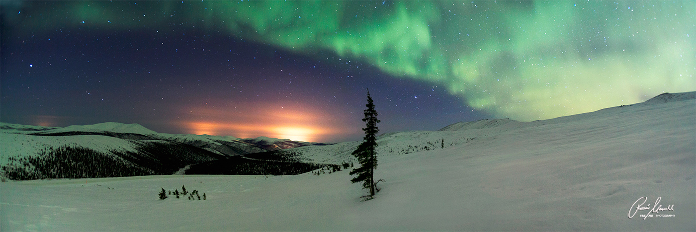 A panoramic image of the green Northern Lights near Fairbanks, Alaska with the orange city lights in the distance